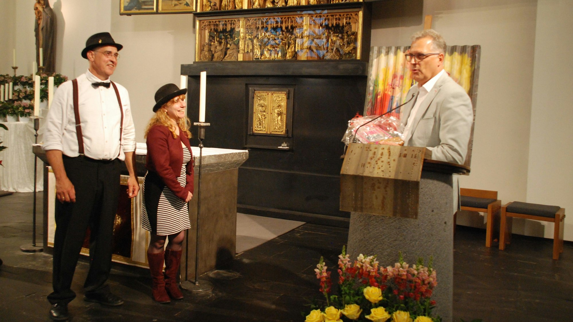 Am Rednerpult in einer Kirche steht Zülpichs Bürgermeister Ulf Hürtgen. Er hält ein Geschenk zum Heimatpreis in der Hand, den er Toni Schleifer und Eugenia Fabrizi verleihen wird. Die beiden stehen links neben ihm.