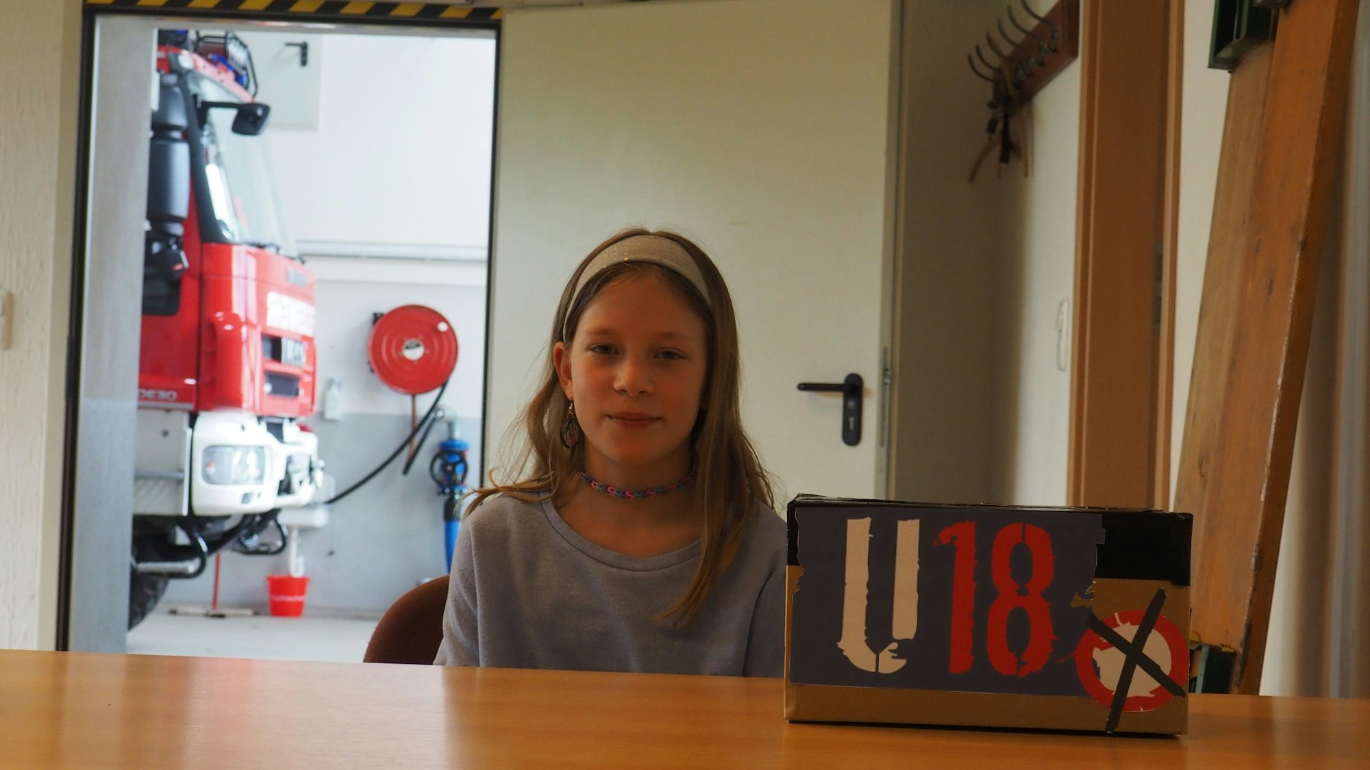 Die Elfjährige sitzt vor ihrer selbstgebastelten Wahlurne mit der Aufschrift U18, im Hintergrund ein Löschfahrzeug der Feuerwehr.