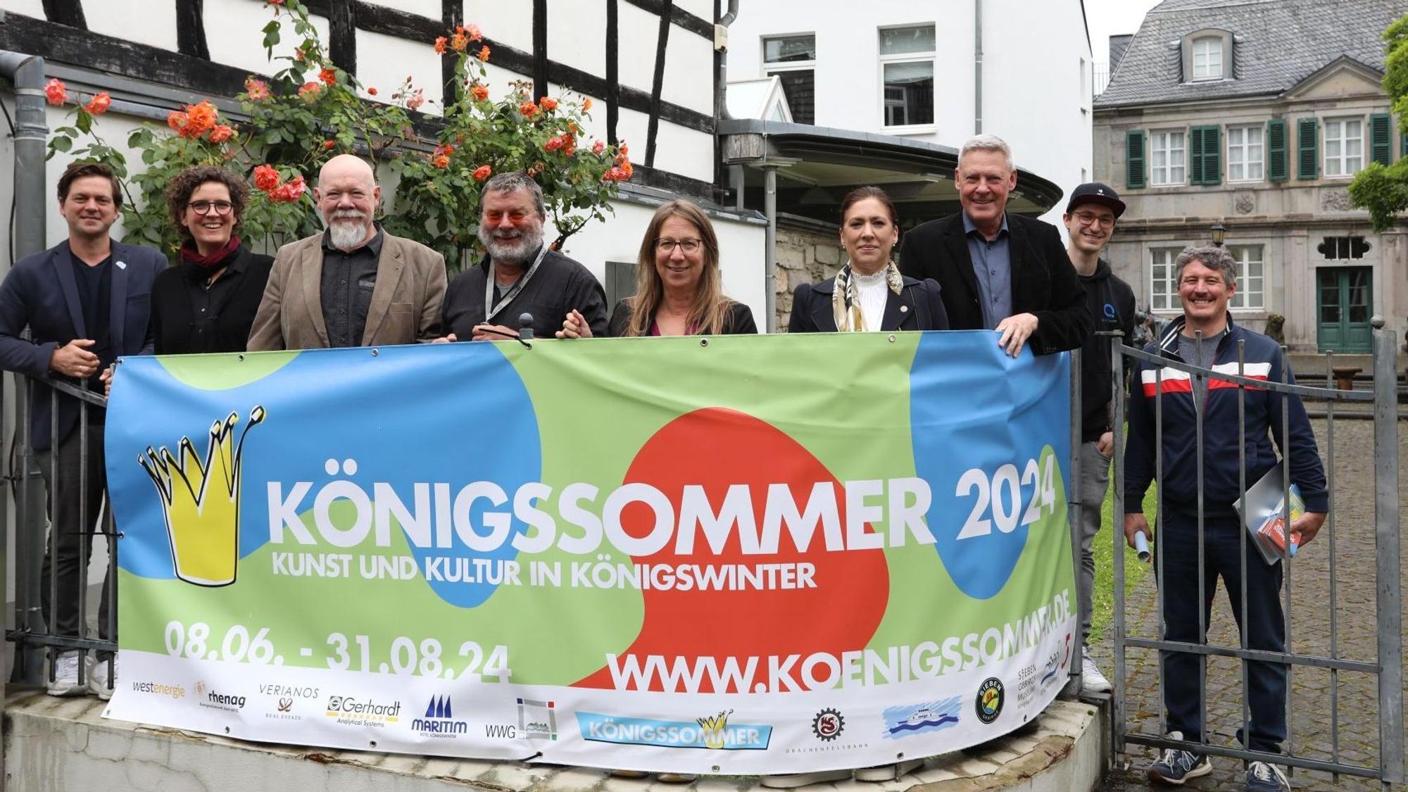 Eine Gruppe von Männern und Frauen stehen hinter einem Transparent mit der Aufschrift „Königssommer 2024“.