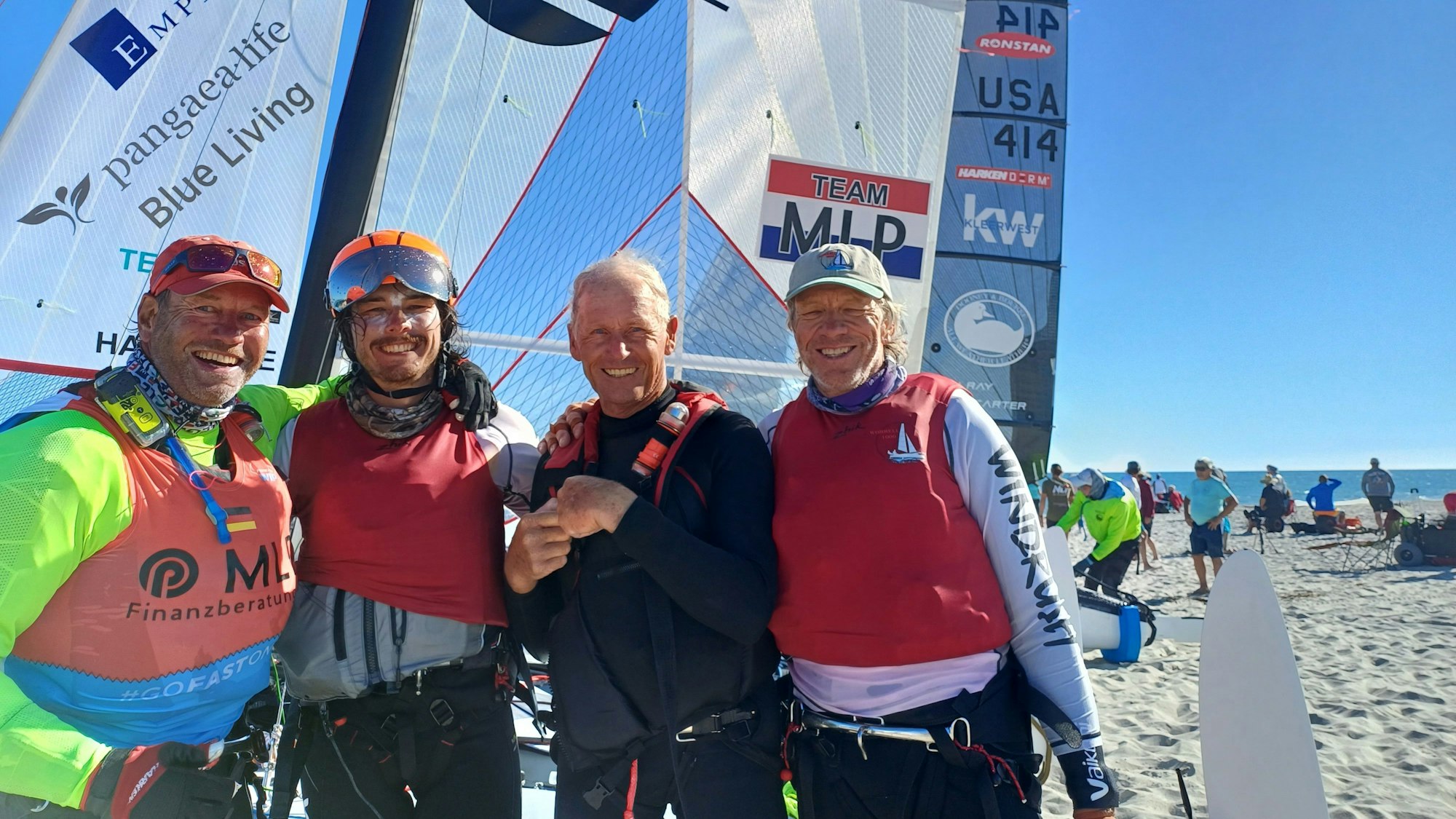 Der Euskirchener André Hauschke ist links im Bild zu sehen. Er posiert mit dem Team Australia 2 und Team MLP am Strand vor einem aufgespannten Segel.