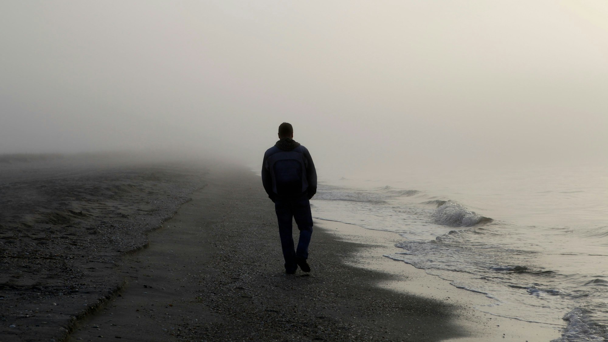 Ein Mann, von dem nur die Umrisse zu erkennen sind, spaziert an einem nebeligen Strand entlang. (Symbolbild)