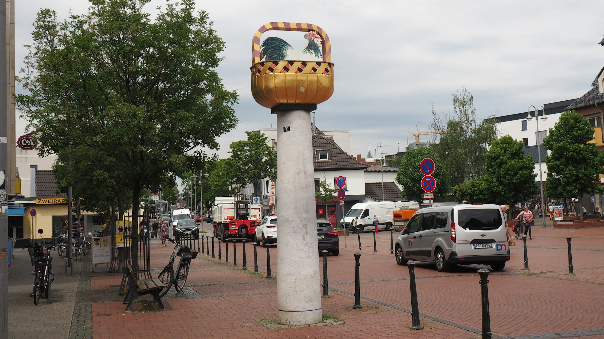 Zu sehen ist eine Säule, auf der ein goldfarbener Korb, befestigt ist. Darin sitzt ein Hahn.