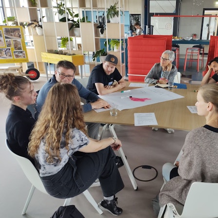 Das Foto zeigt eine der sieben Arbeitsgruppen, deren Mitglieder an einem Tisch Platz genommen haben. In der Mitte liegt ein großes Blatt, auf das die Gedanken und Ergebnisse geschrieben wurden.