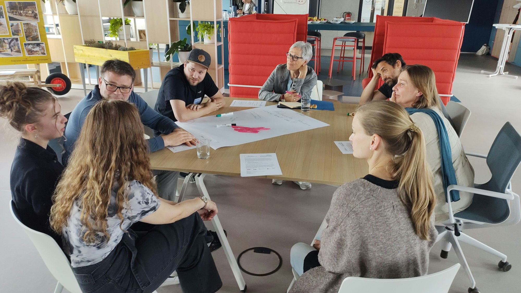 Das Foto zeigt eine der sieben Arbeitsgruppen, deren Mitglieder an einem Tisch Platz genommen haben. In der Mitte liegt ein großes Blatt, auf das die Gedanken und Ergebnisse geschrieben wurden.