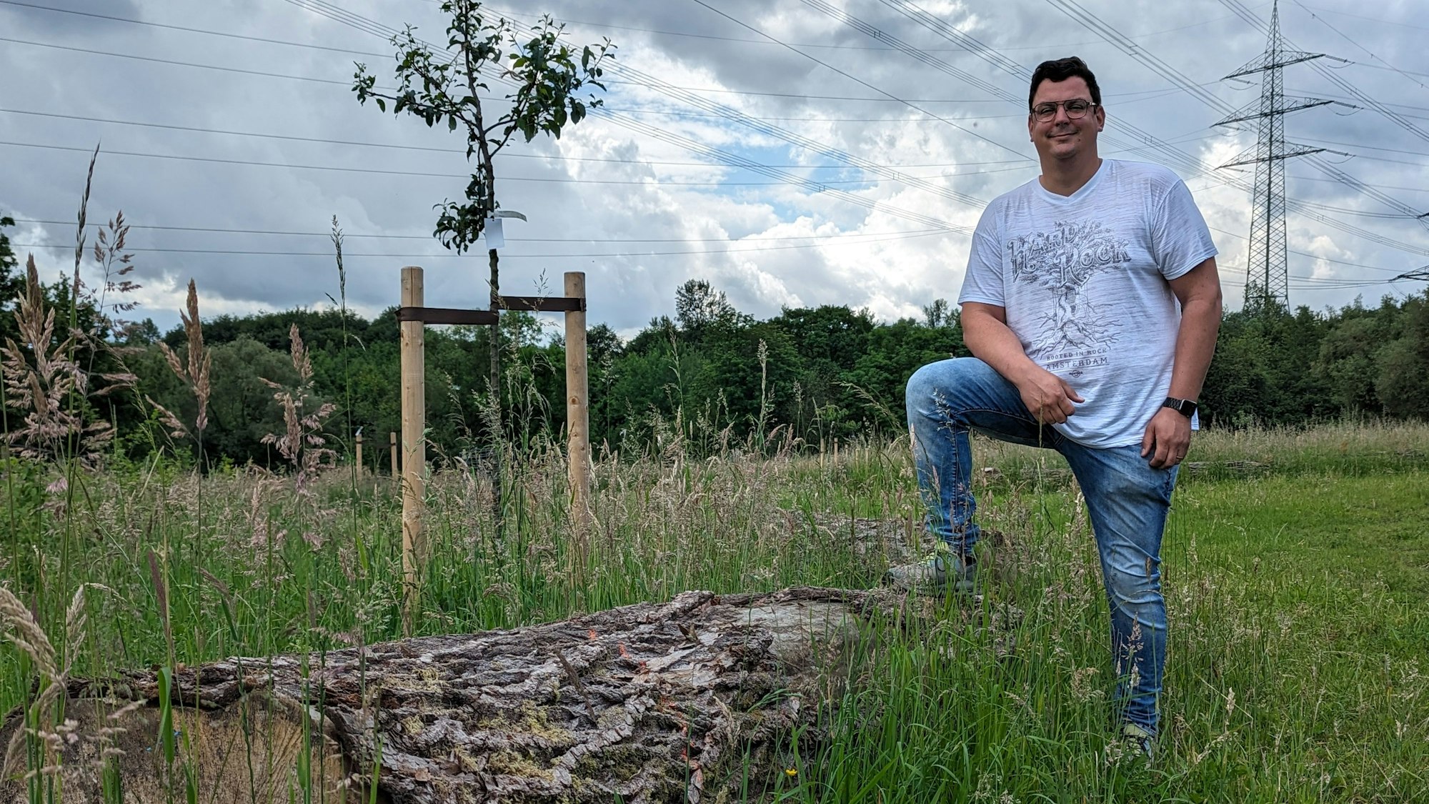 Ein Mann steht auf einer Wiese mit frisch gepflanzten Bäumen, ein Bein auf einen alten Baumstamm gestellt.