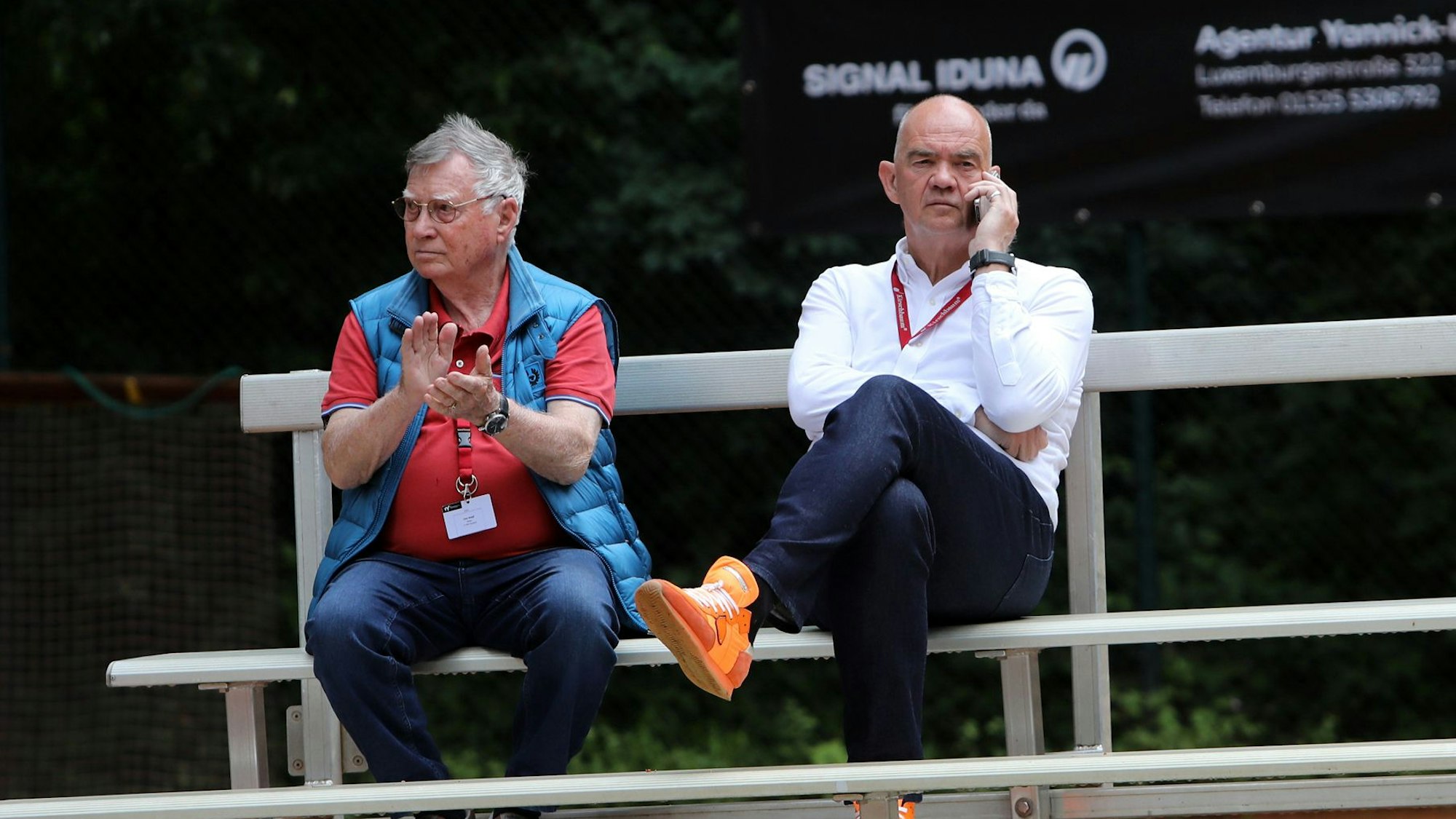 Zwei Männer sitzen auf eine Bank, einer telefoniert, einer applaudiert.