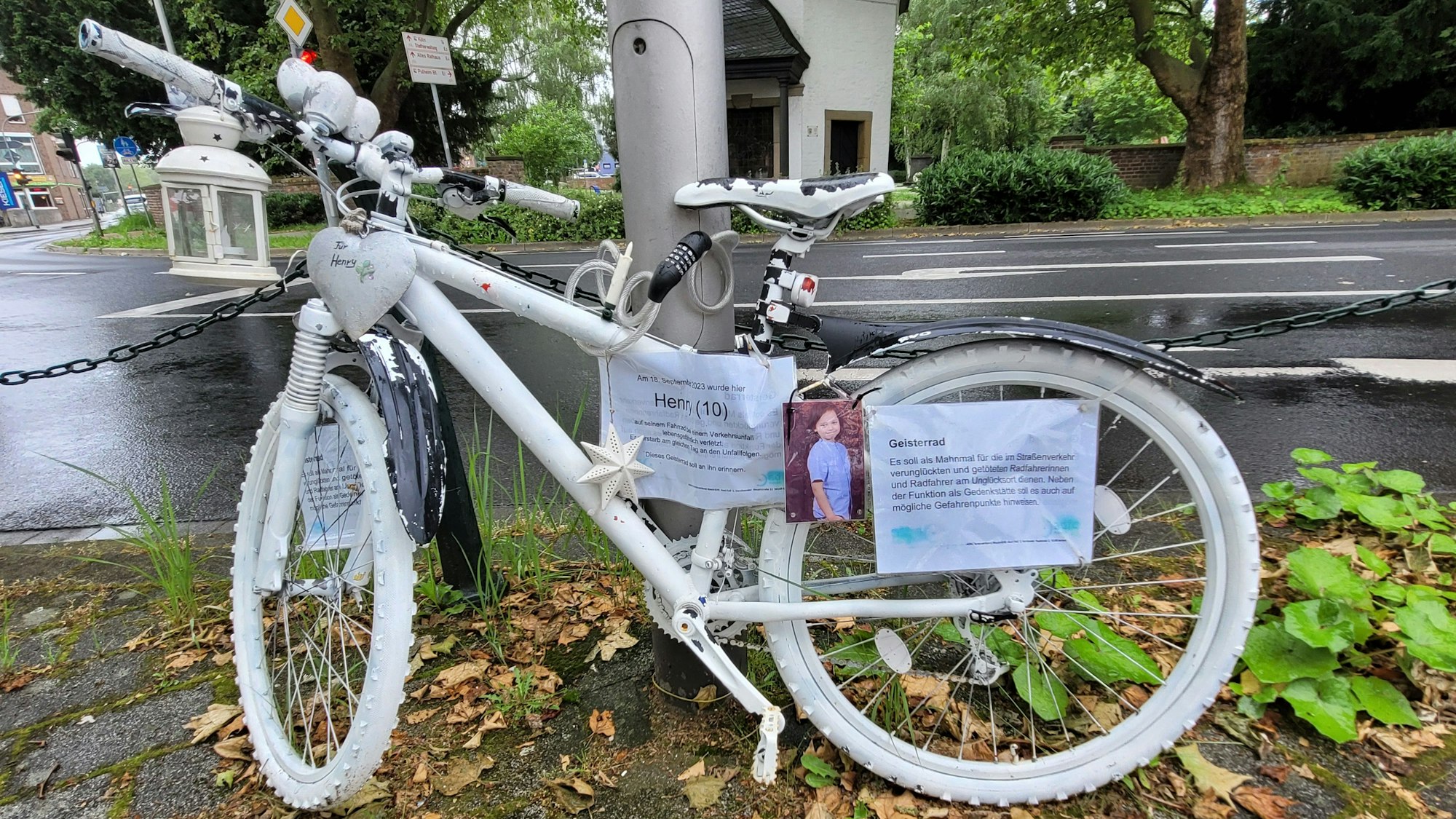 Ein weiß-lackiertes Fahrrad, es steht an der Unfallstelle. Daran befestigt ist ein Foto des verstorbenen Jungen.