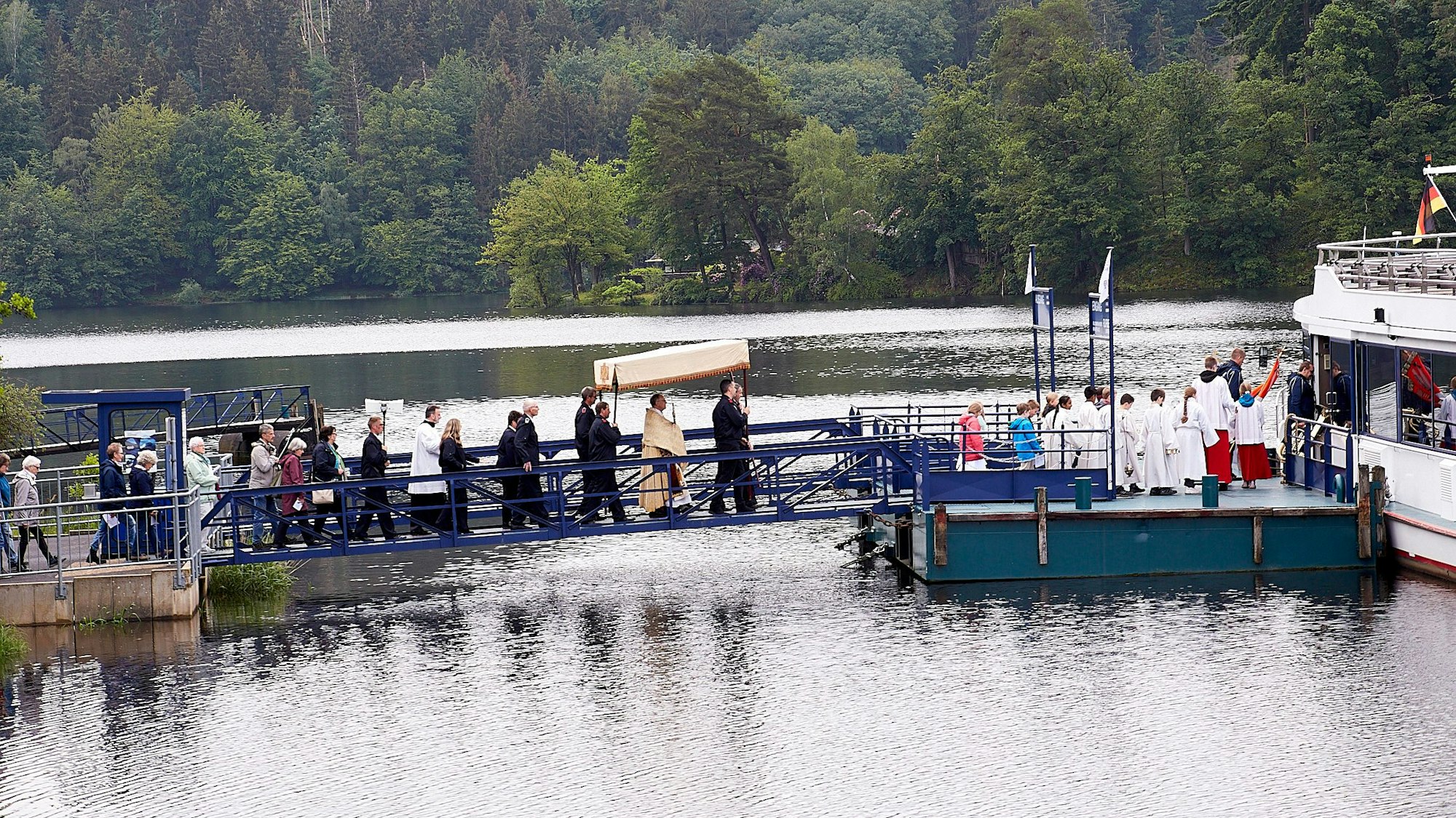 Eine Prozession zieht über einen Anleger am Rursee auf ein Schiff. In der Mitte ist der Priester zu sehen, der unter einem Baldachin geht und eine Monstranz trägt.