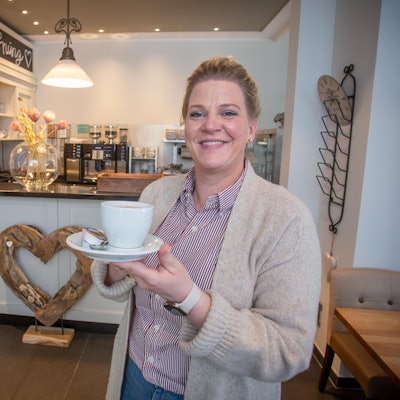Eine Frau schaut in die Kamera, sie hat eine Tasse Kaffee mit Untertasse i der Hand, im Hintergrund sind ein Tresen und eine Kaffeemaschine zu sehen. Auf einer Tafel im oberen Bildrand steht das Wort „Neueröffnung“.