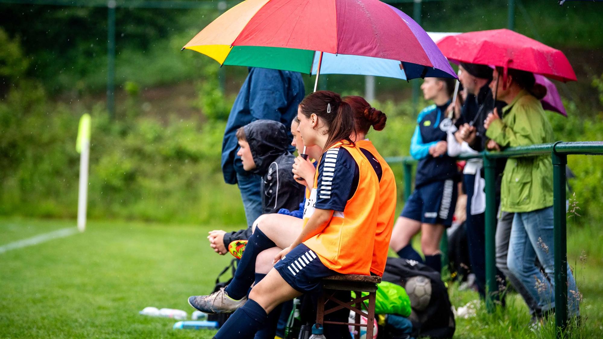 Das Bild zeigt die Auswechselbank des VfL Kommern. Einige Spielerinnen sitzen unter einem bunten Regenschirm.