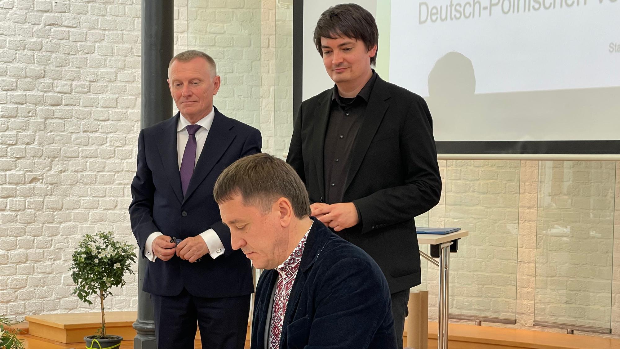 Ein man sitzt an einem Tisch und unterzeichnet ein Papier, hinter ihm stehen zwei weitere Männer im Anzug.