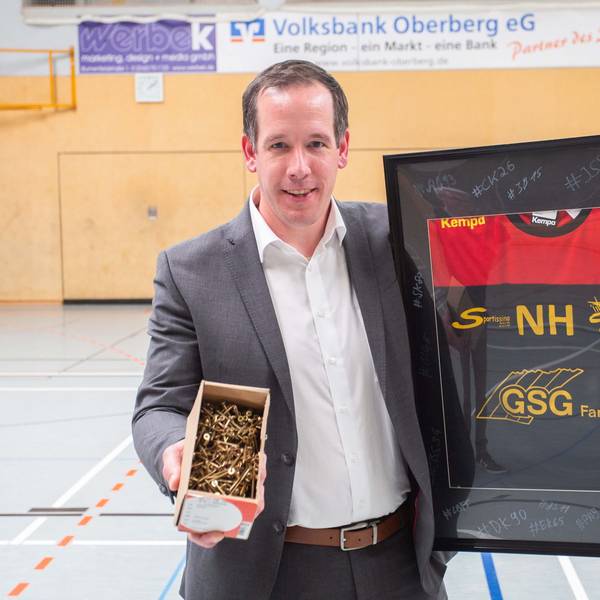 Nils Hühn hält ein eingerahmtes Handballtrikot und eine Koste voller Schrauben in den Händen. Er steht in einer Sporthalle.