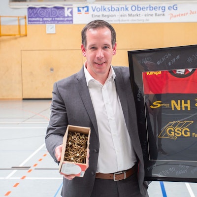 Nils Hühn hält ein eingerahmtes Handballtrikot und eine Koste voller Schrauben in den Händen. Er steht in einer Sporthalle.