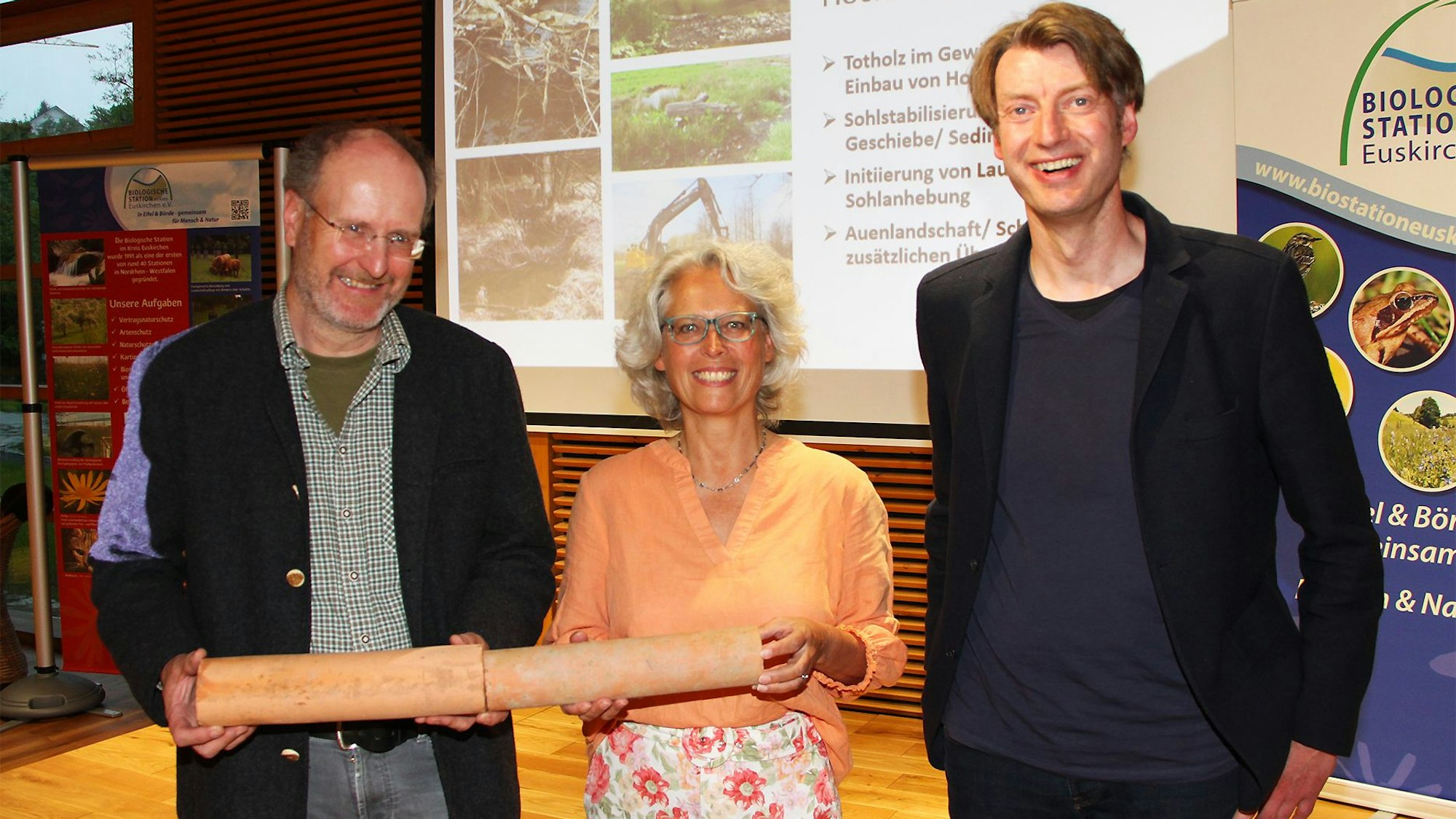 Zwei Männer und eine Frau stehen im Holzkompetenzzentrum in Nettersheim. Zwei halten Tonrohre fest, die als Drainagen in Weisen verwendet werden.