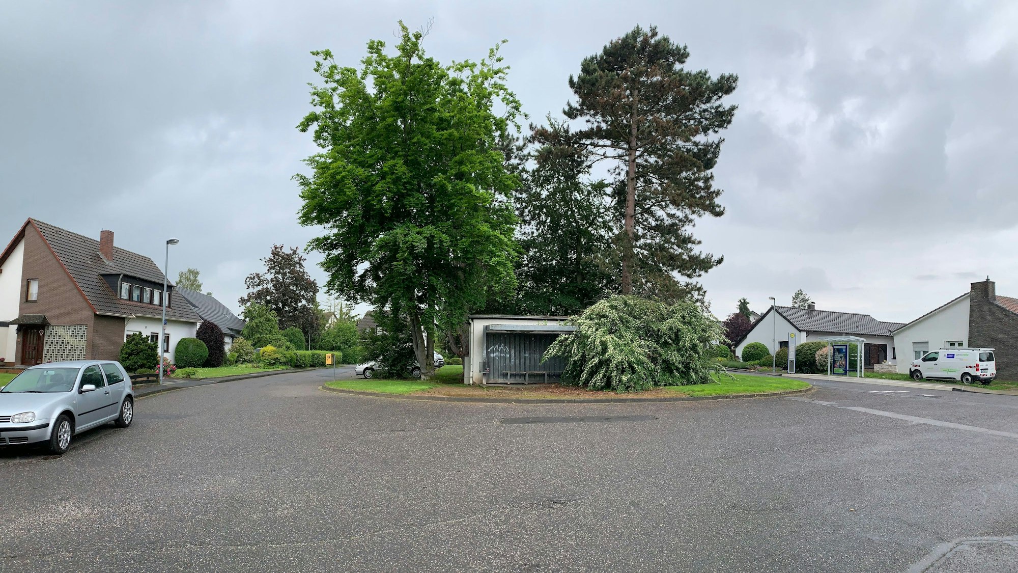 Auf dem Platz stehen Bäume, ein altes Buswartehäuschen und Autos. Er ist von Straßen mit Einfamilienhäusern gesäumt.