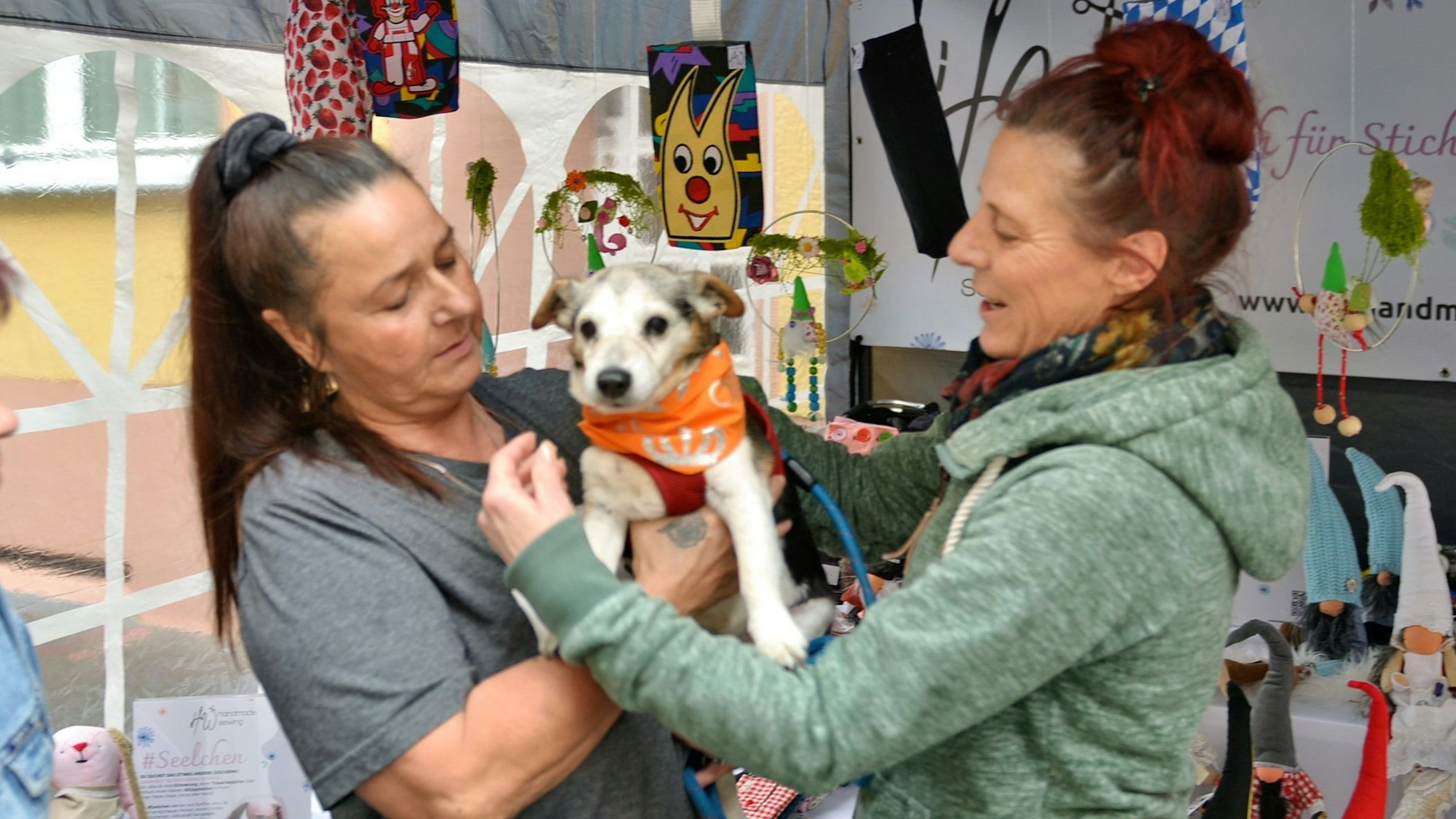Eine Frau hält ihren kleinen Hund auf dem Arm, eine andere legt ihm ein Halstuch an.
