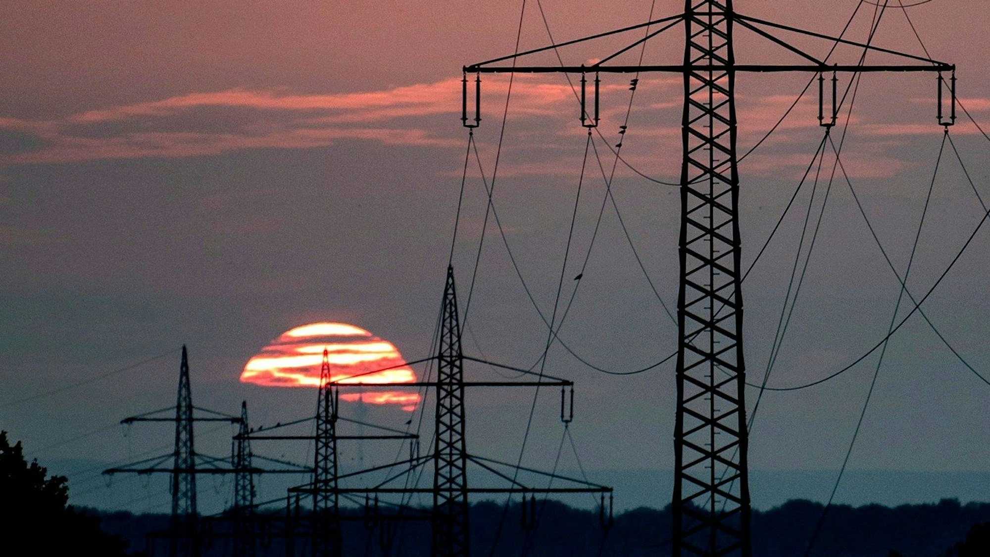 Im Sonnenuntergang sind einige Strommasten und Leitungen zu sehen. Die Sonne hat die Wolken rosa gefärbt.