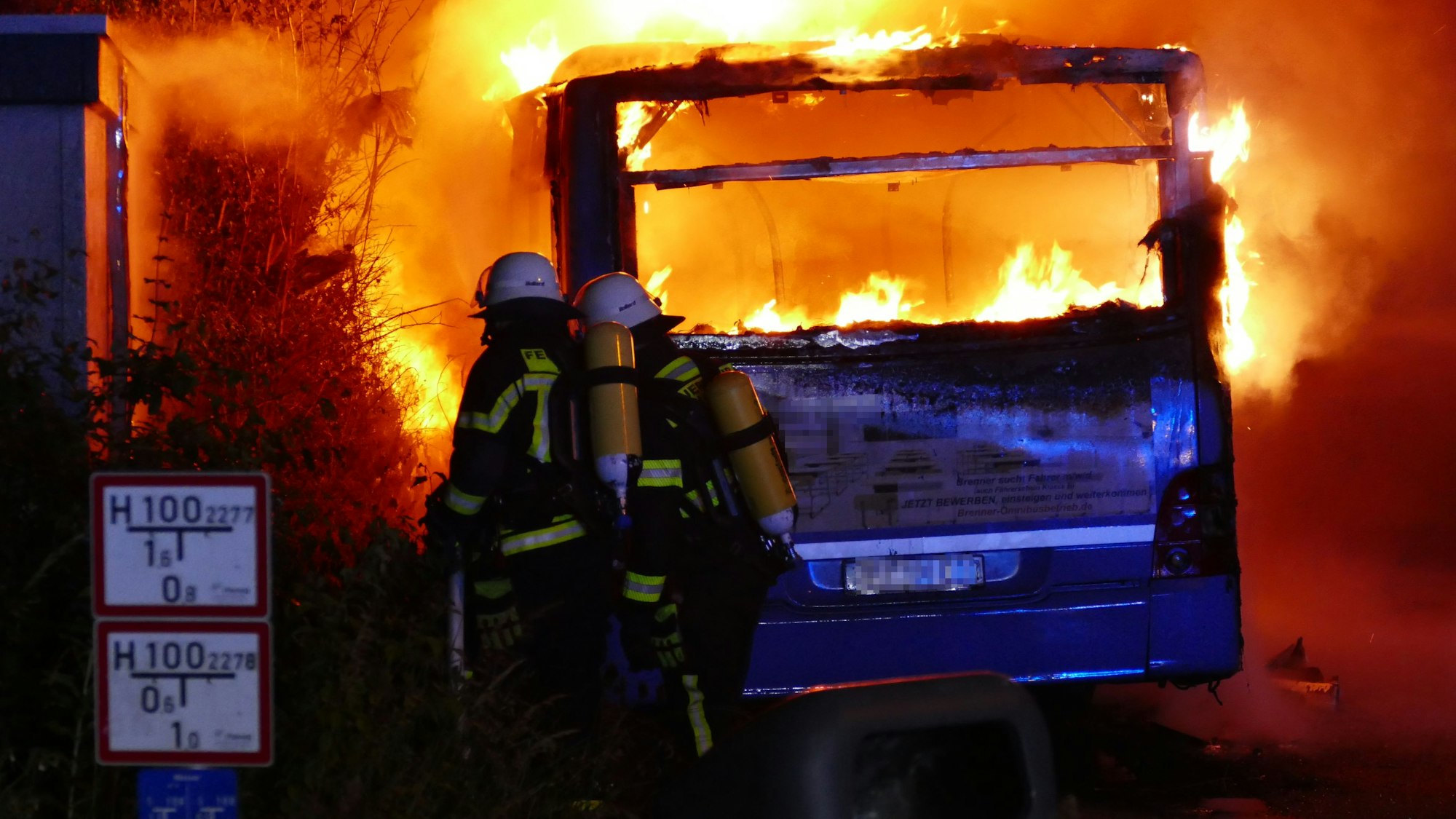 Feuerwehrleute stehen hinter einem brennenden Linienbus.