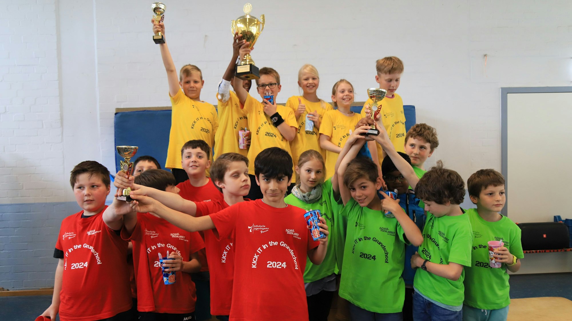 Kinder in roten, grünen und gelben T-Shirts bei einem Gruppenfoto.Sie recfken Pokale nach oben.