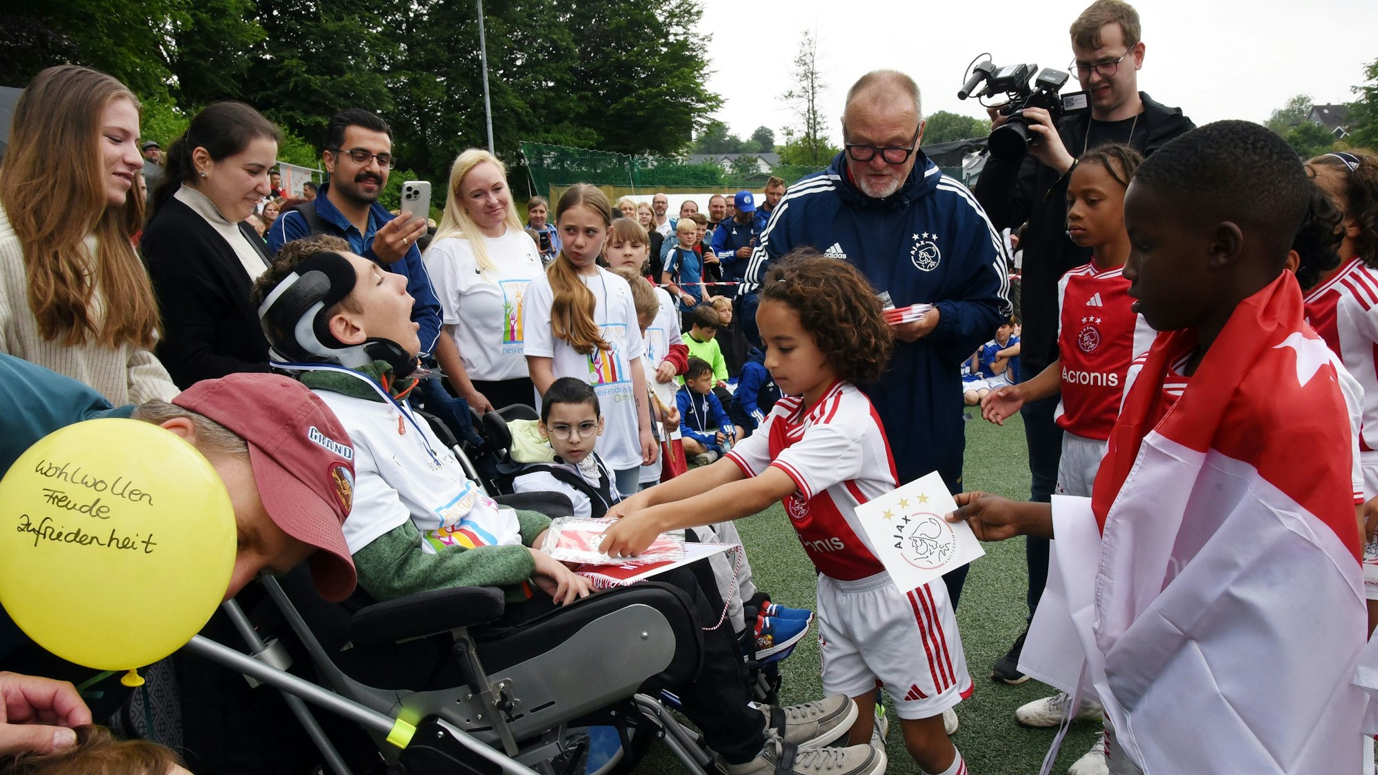 Die U19-Fußballer von Ajax Amsterdam schauen zu, wir ein Vereinskollege an einen Jungen im Rollstuhl einen Vereinswimpel verschenkt.