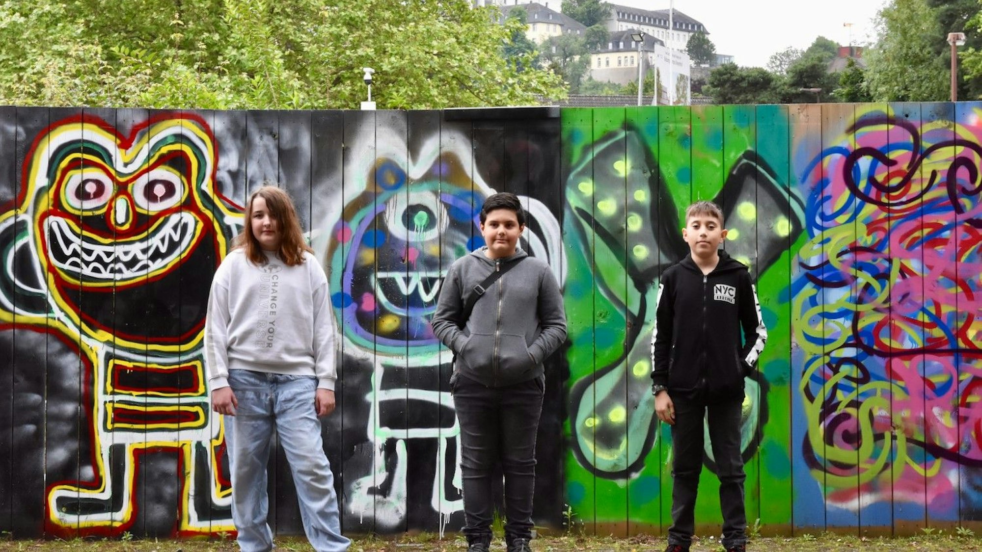 Zwei Jungen und ein Mädchen stehen vor einem Bauzaun, der mit bunten Graffiti bedeckt ist. im Hintergrund ist eine Klosterkirche auf einem Berg zu sehen.