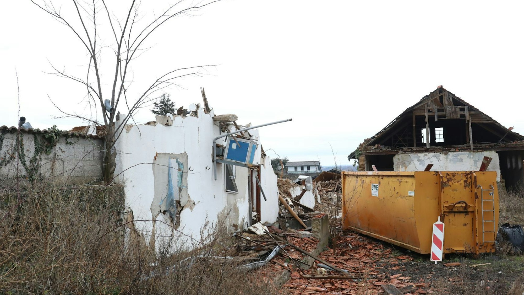 Ein orangefarbener Baucontainer steht neben Resten einer Gebäudefassade und einer teilweise abgerissenen Scheune.