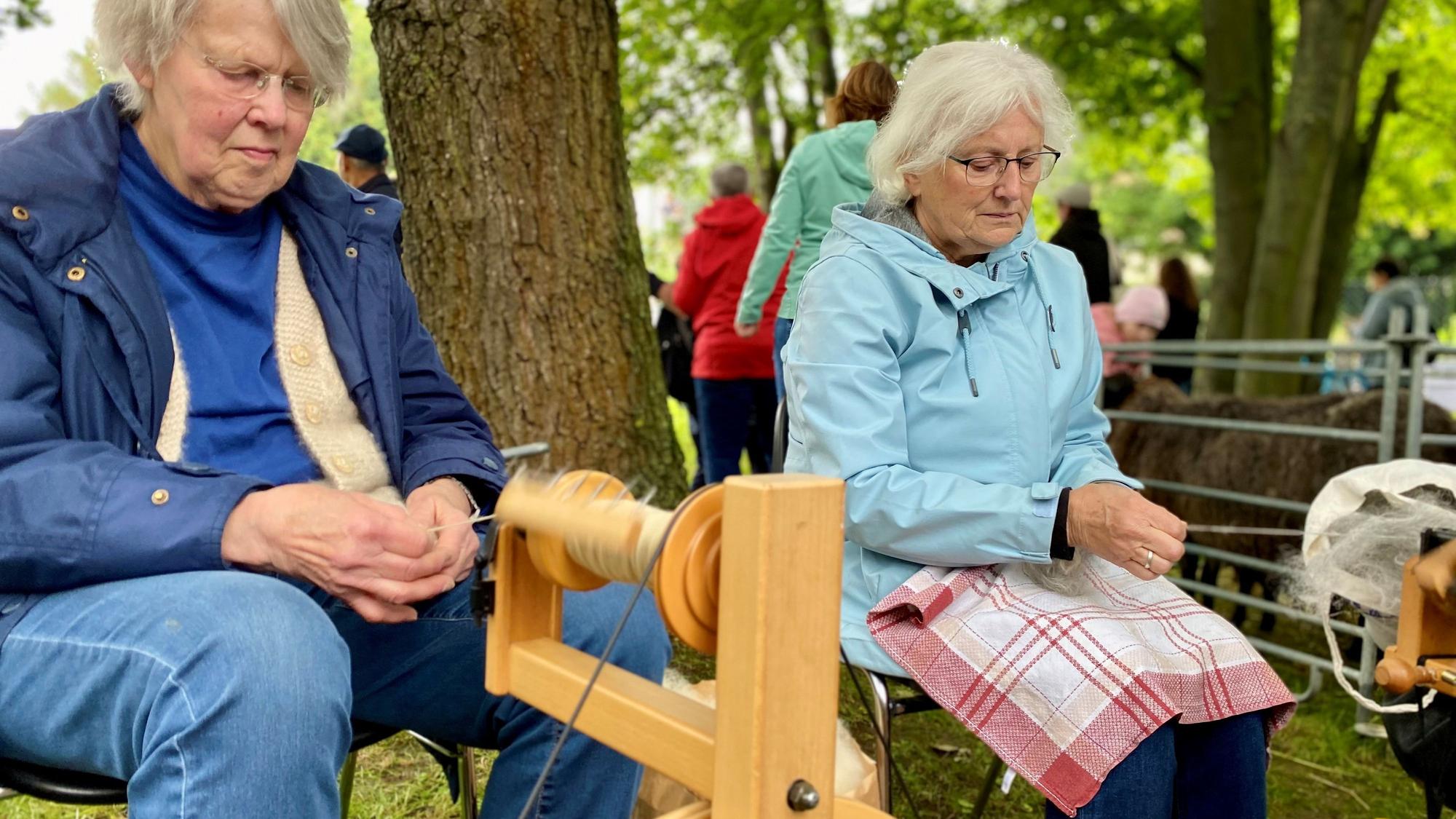 Ursula Metten und Maria Lieven sitzen an zwei Spinnrädern aus Holz.