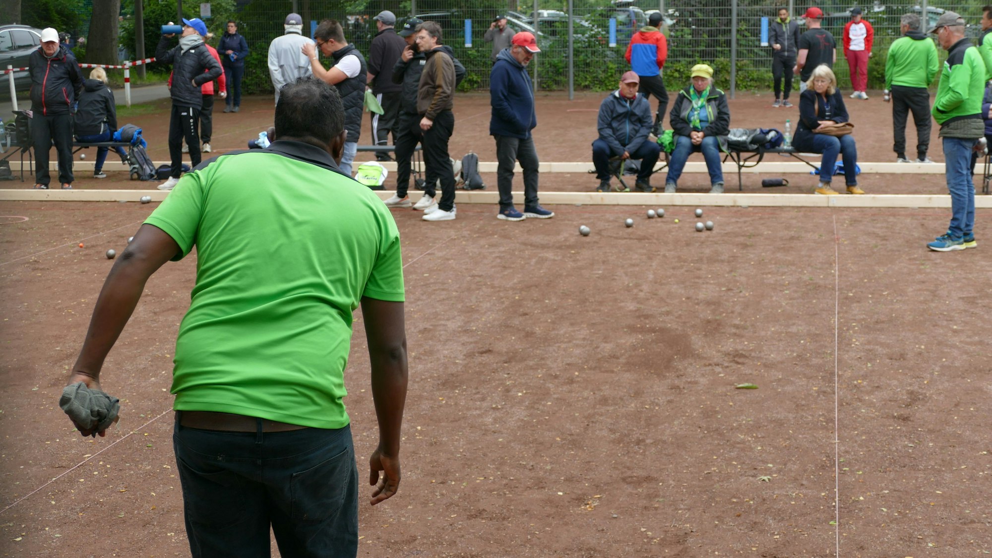 Ein Mann im grünen T-Shirt wirft eine Boulekugel. Im Hintergrund schauen Mitspieler zu.