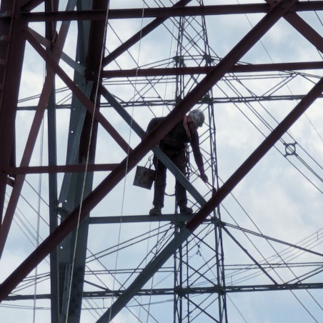Ein Arbeiter mit Farbeimer und Pinseln streicht eine Strebe hoch oben in einem Stahlgittermast.