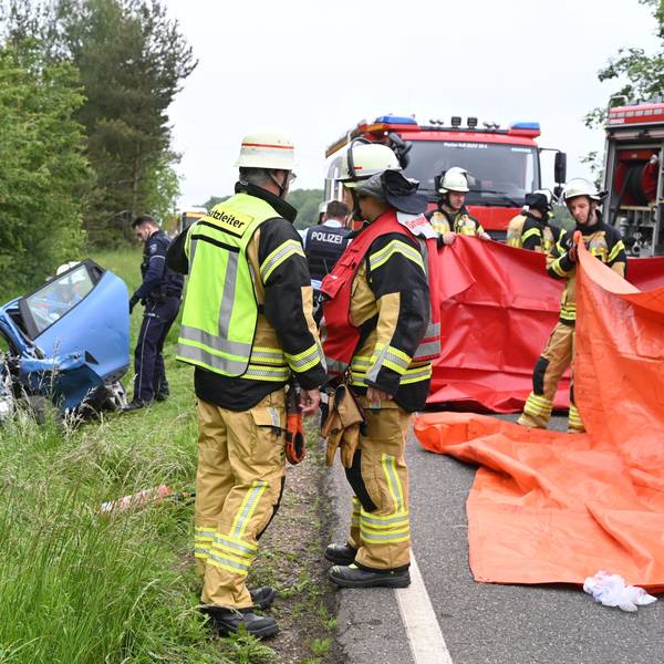 Ein blaues Auto liegt links im Straßengraben, der Fahrer ist bei dem Unfall ums Leben gekommen. Rechts sind Feuerwehrleute zu sehen, die rote Planen als Sichtschutz hochhalten.