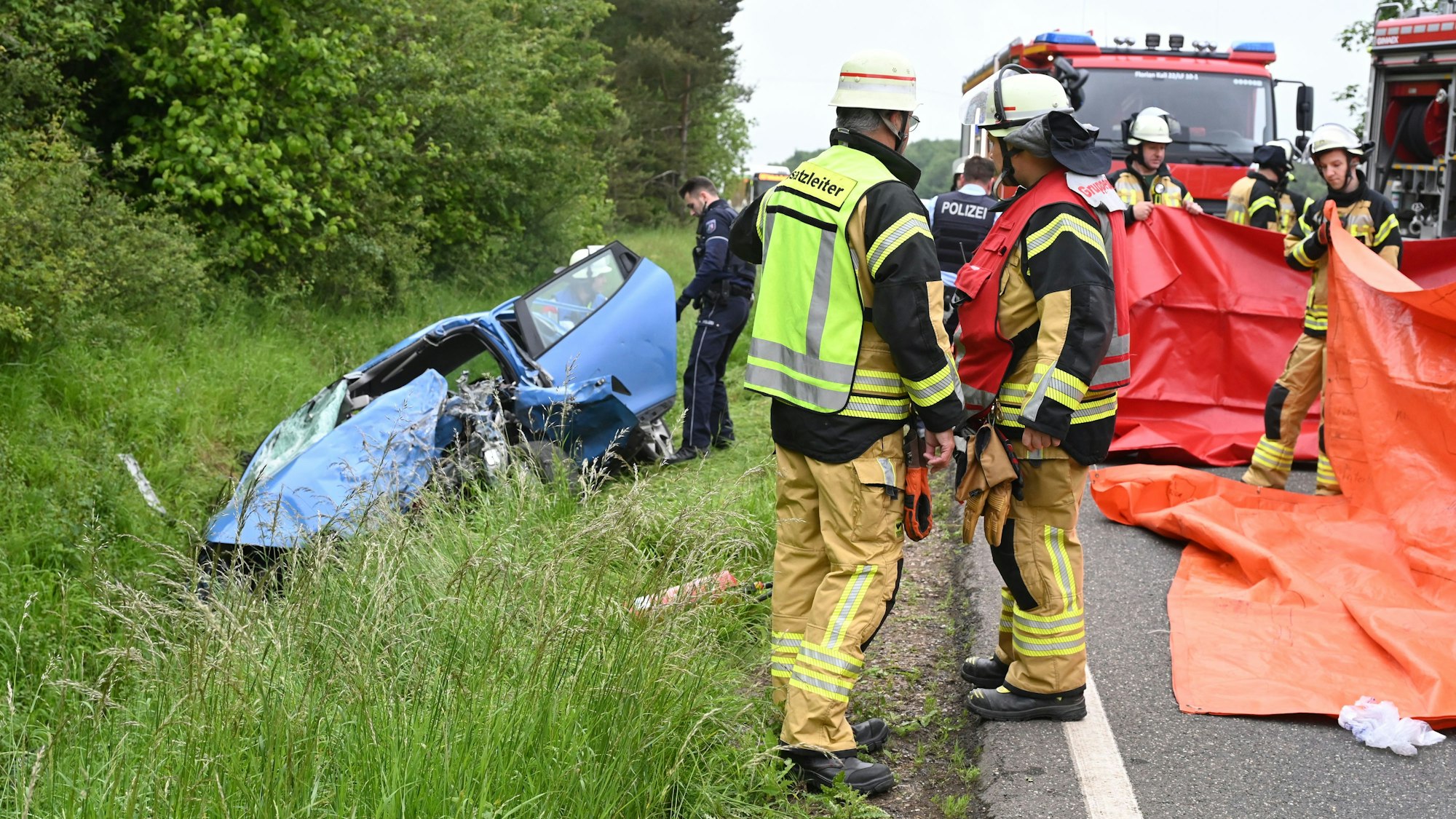 Ein blaues Auto liegt links im Straßengraben, der Fahrer ist bei dem Unfall ums Leben gekommen. Rechts sind Feuerwehrleute zu sehen, die rote Planen als Sichtschutz hochhalten.