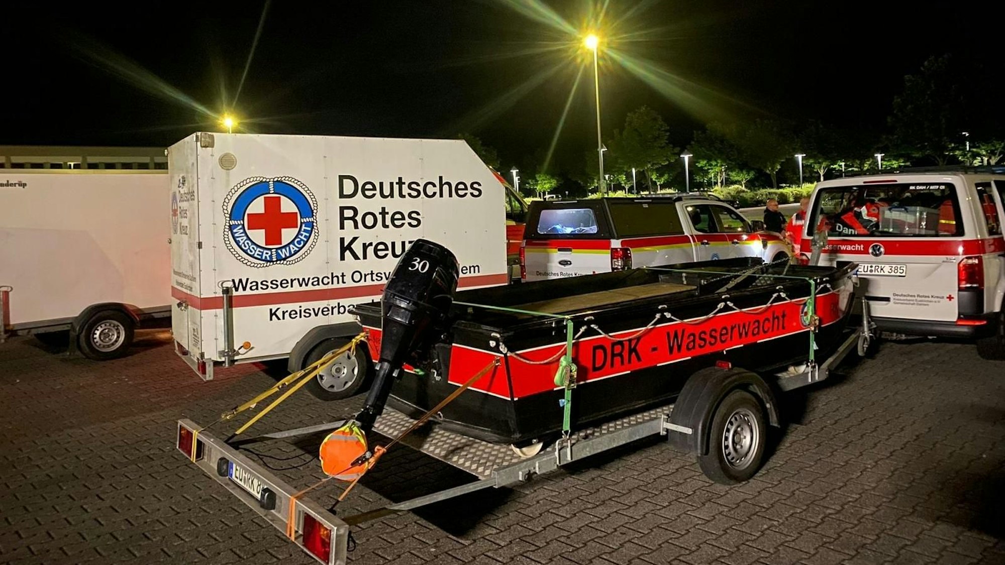 Mehrere Fahrzeuge des Deutschen Roten Kreuzes samt Anhängern stehen in der Nacht auf einem beleuchteten Parkplatz. Auf einem Anhänger ist ein schwarz-rotes Boot mit der Aufschrift „DRK – Wasserwacht“ verladen.
