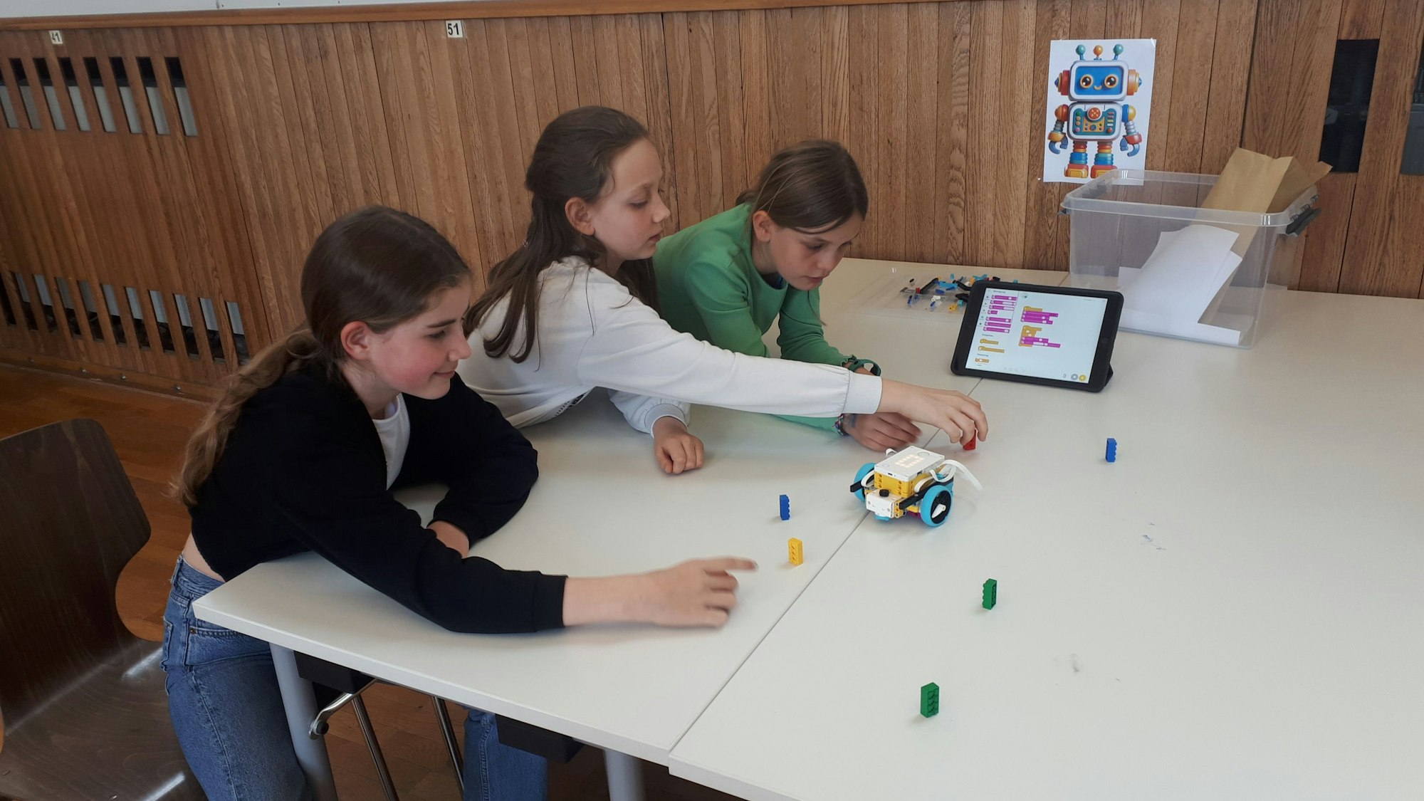 Drei Schülerinnen sitzen an einem weißen Tisch und führen gemeinsam das Funktionsprinzip eines Lego-Roboters vor. Foto von Bernd Schöneck