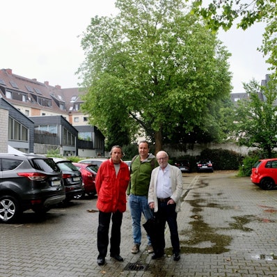 Die Bezirksvertreter Roland Schüler, Florian Weber-Baronowsky (Grüne) und Friedhelm Hilgers stehen auf dem Parkplatz an der Redwitzstraße 62 in Köln-Sülz und vor der großen Platane im Hintergrund. Foto von Susanne Esch.