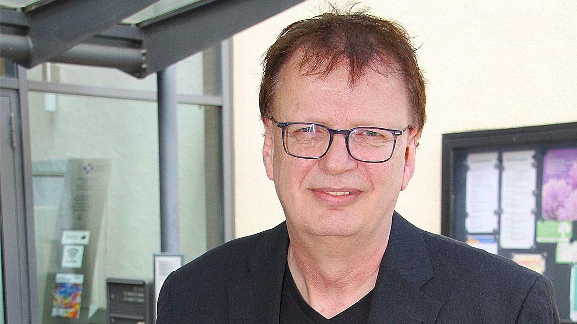 Das Porträt zeigt Jens Schramm. Er steht im Eingangsbereich eines Gebäudes in Euskirchen, im Hintergrund ist ein Schaukasten mit mehreren Informationsblättern zu sehen.