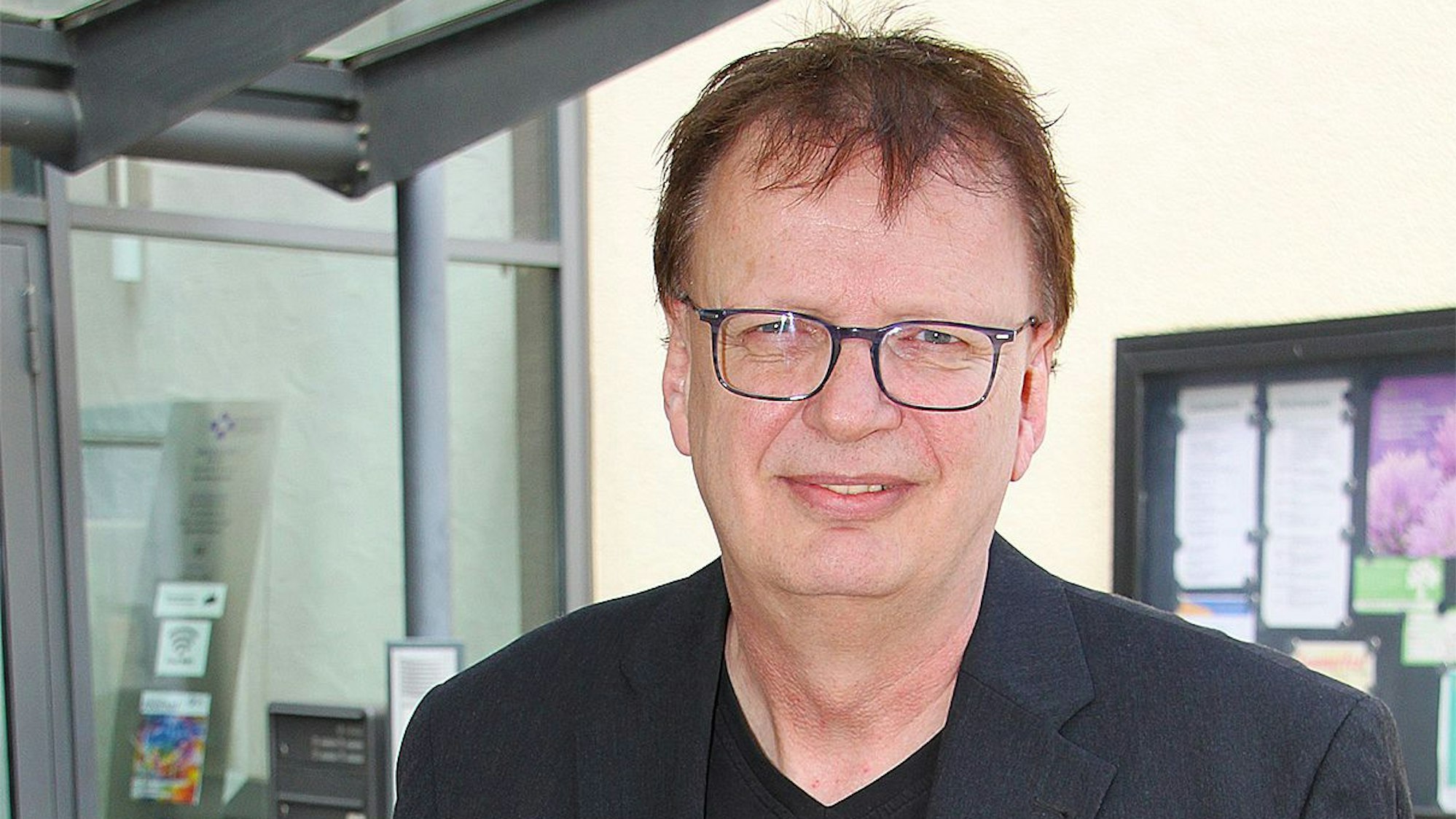 Das Porträt zeigt Jens Schramm. Er steht im Eingangsbereich eines Gebäudes in Euskirchen, im Hintergrund ist ein Schaukasten mit mehreren Informationsblättern zu sehen.