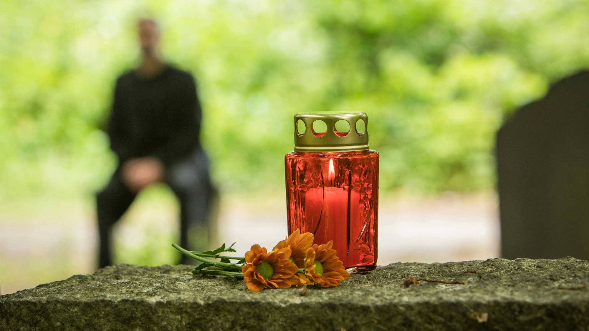 Vor einem roten Grablicht auf einem Stein liegen einzelne Blumen. Dahinter ist unscharf ein sitzender Mann zu sehen.