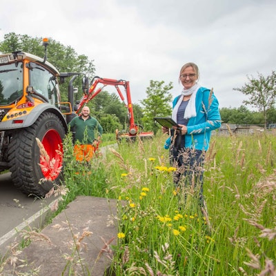 Manuela Thomas steht mit einem Tablet an einem dicht bewachsenen Wegesrand. Hinter ihr steht Christoph Seelbach neben einer großen Nähmaschine.
