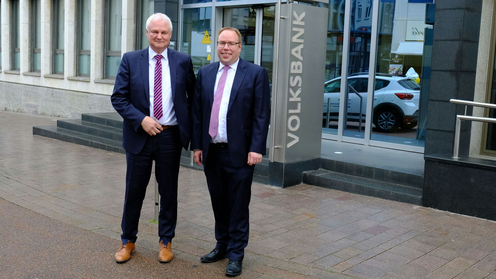 Die beiden Vorstandsmitglieder der Volksbank Euskirchen, Hans-Jürgen Lembicz (l.) und Marc Güttes, stehen vor dem Haupteingang der Bank.
