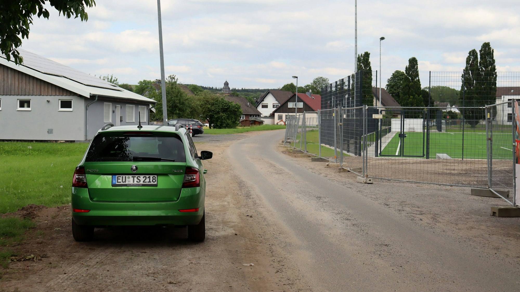 Die Straße im Floting. Vorne parkt ein Auto, rechts ist der neue Kunstrasenplatz zu erkennen, links das Heim des Musikvereins Eifelland.