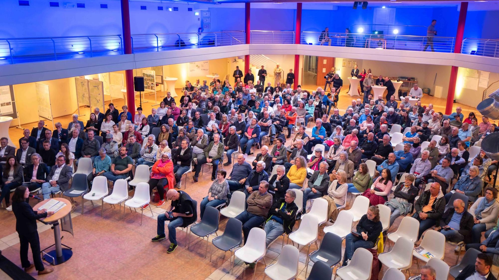 Zahlreiche Menschen sitzen in einem Saal auf Stühlen. Links steht Landrätin Cornelia Weigand an einem Pult und spricht in ein Mikrofon.