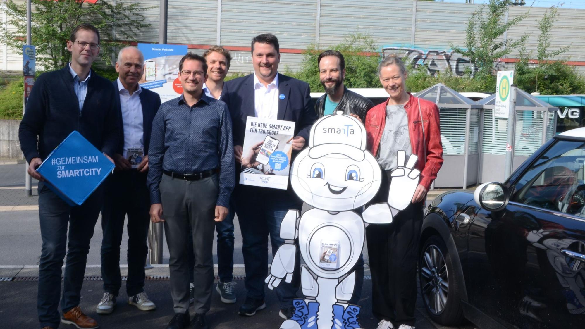 Männer und Frauen stehen auf einem Parkplatz. im Hintergrund ist eine Bushaltestelle zu sehen, vor der Gruppe steht die winkende Pappfigur eines Roboters.