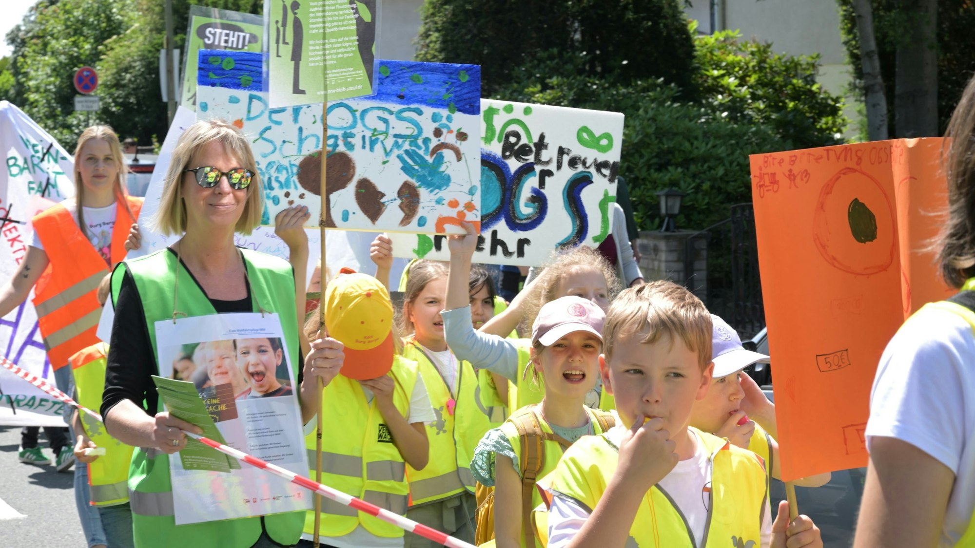 Kinder in gelben Warnwesten und mit Trillerpfeifen ziehen mit Protestplakaten über eine Straße.