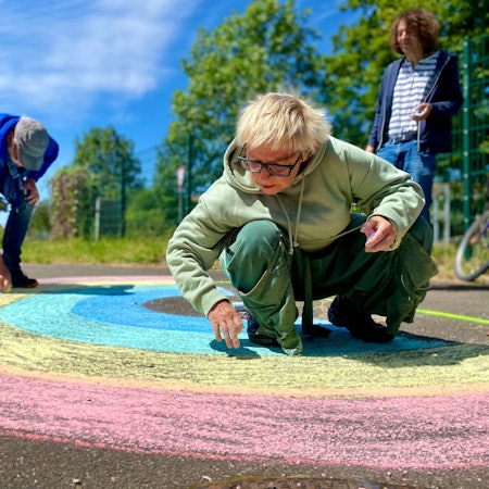 Anne Bergmann, Leo Schmidt und Thomas Latzke malen mit Straßenkreide einen Regenbogen auf die Straße.