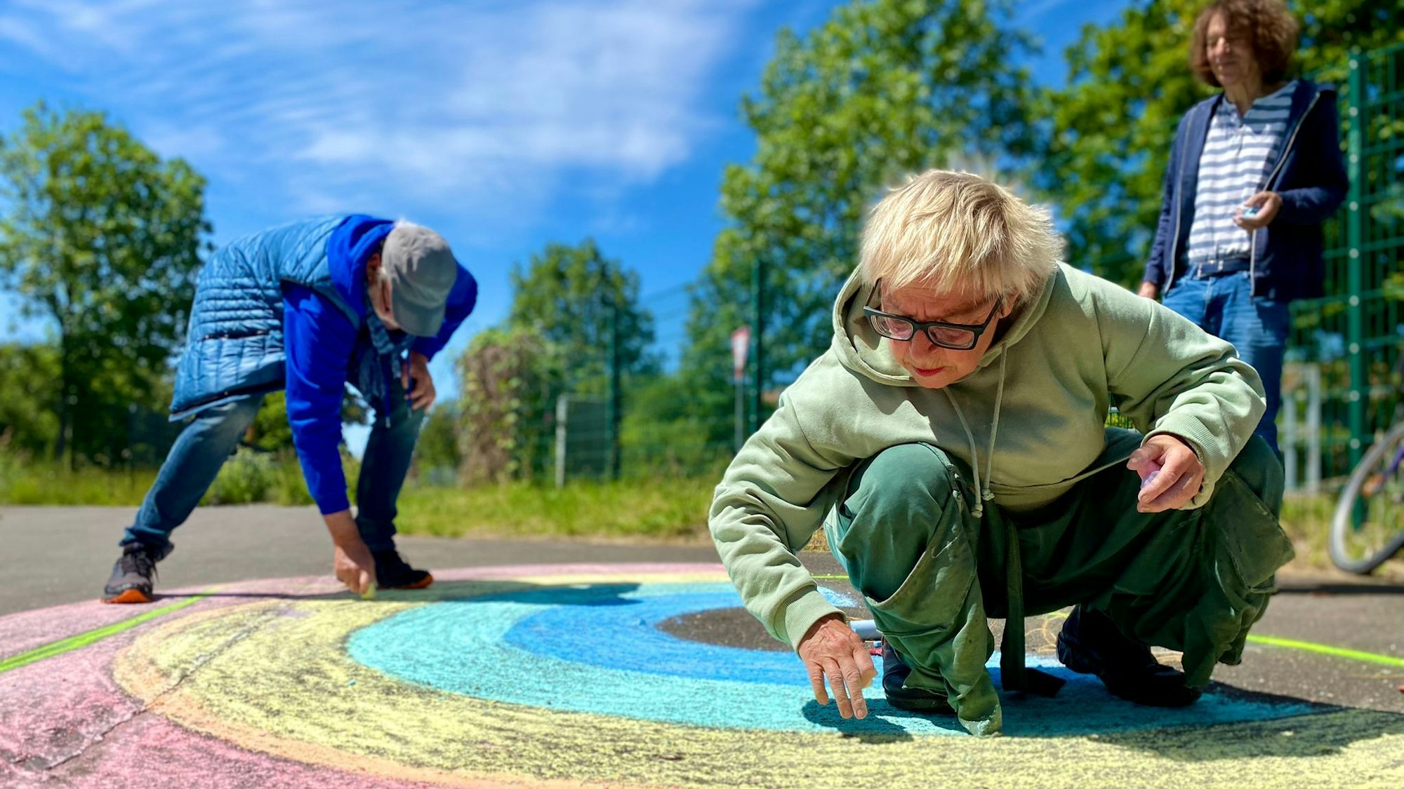 Anne Bergmann, Leo Schmidt und Thomas Latzke malen mit Straßenkreide einen Regenbogen auf die Straße.