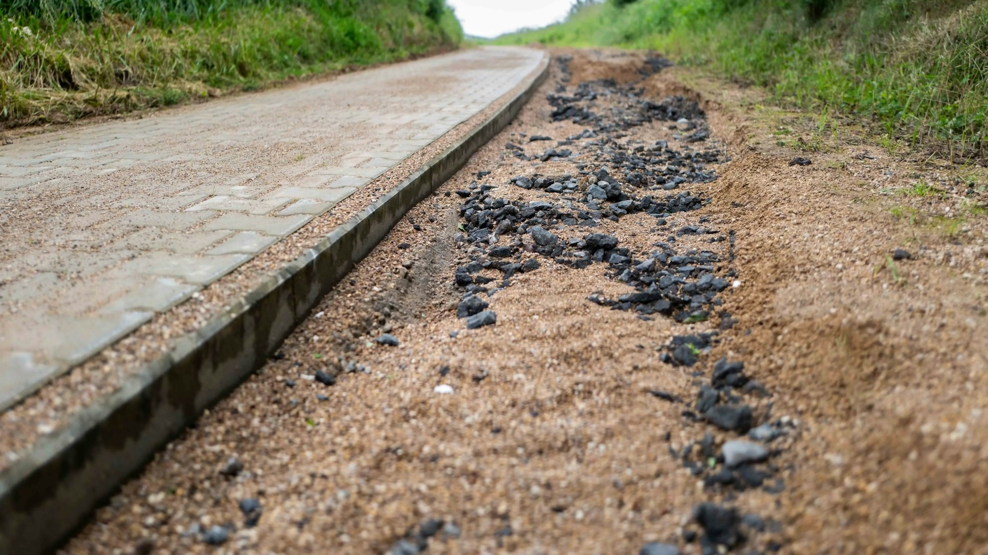 Das Bild zeigt einen recht hohen Versatz zwischen gepflastertem Radweg und dem Fußweg. Kies und Schotter sind durch den Regen teilweise ausgespült.