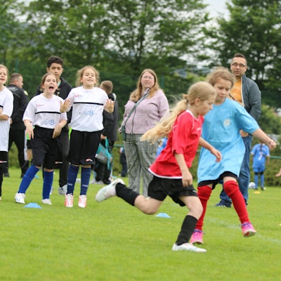 Zwei Mädchen spielen Fußball, Zuschauer stehen am Rand und fiebern mit.