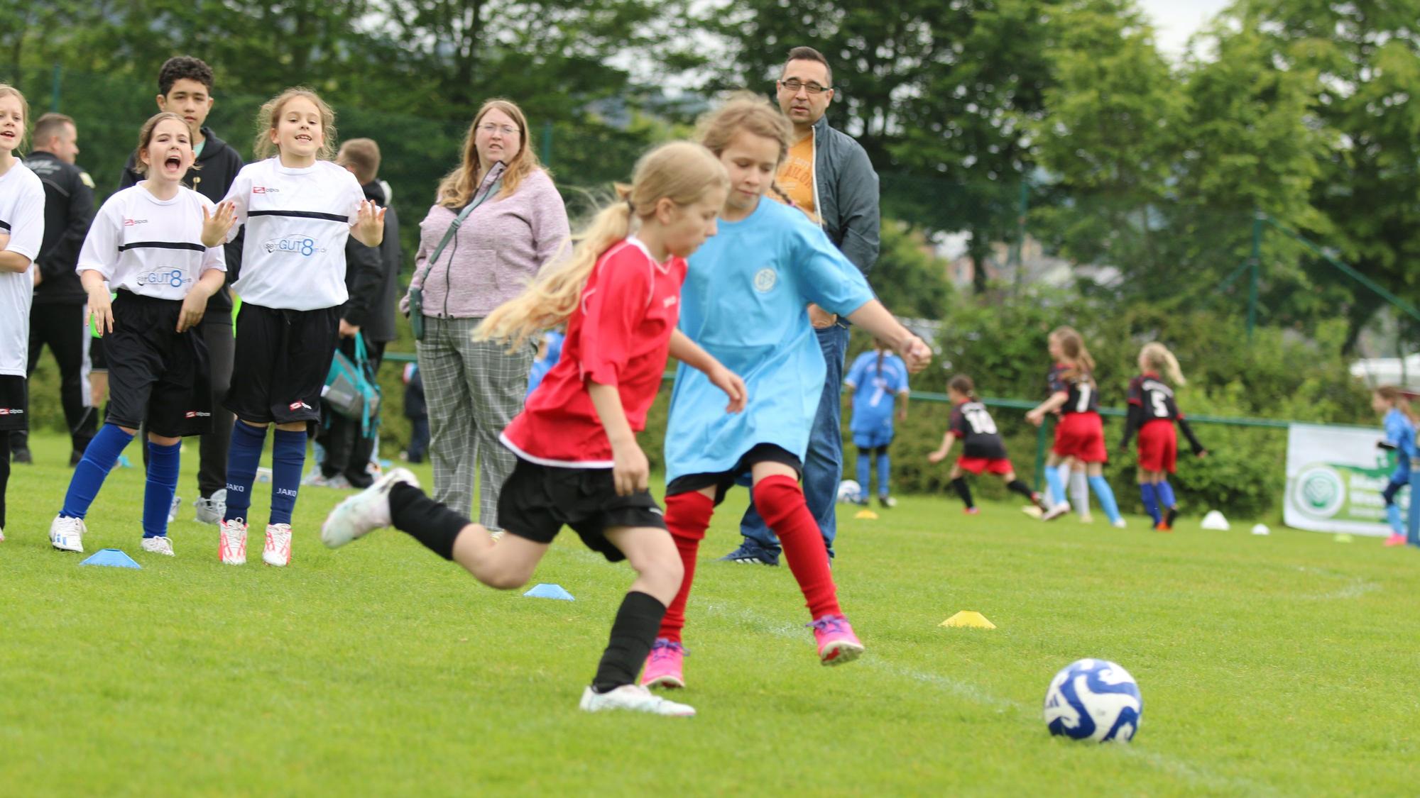 Zwei Mädchen spielen Fußball, Zuschauer stehen am Rand und fiebern mit.