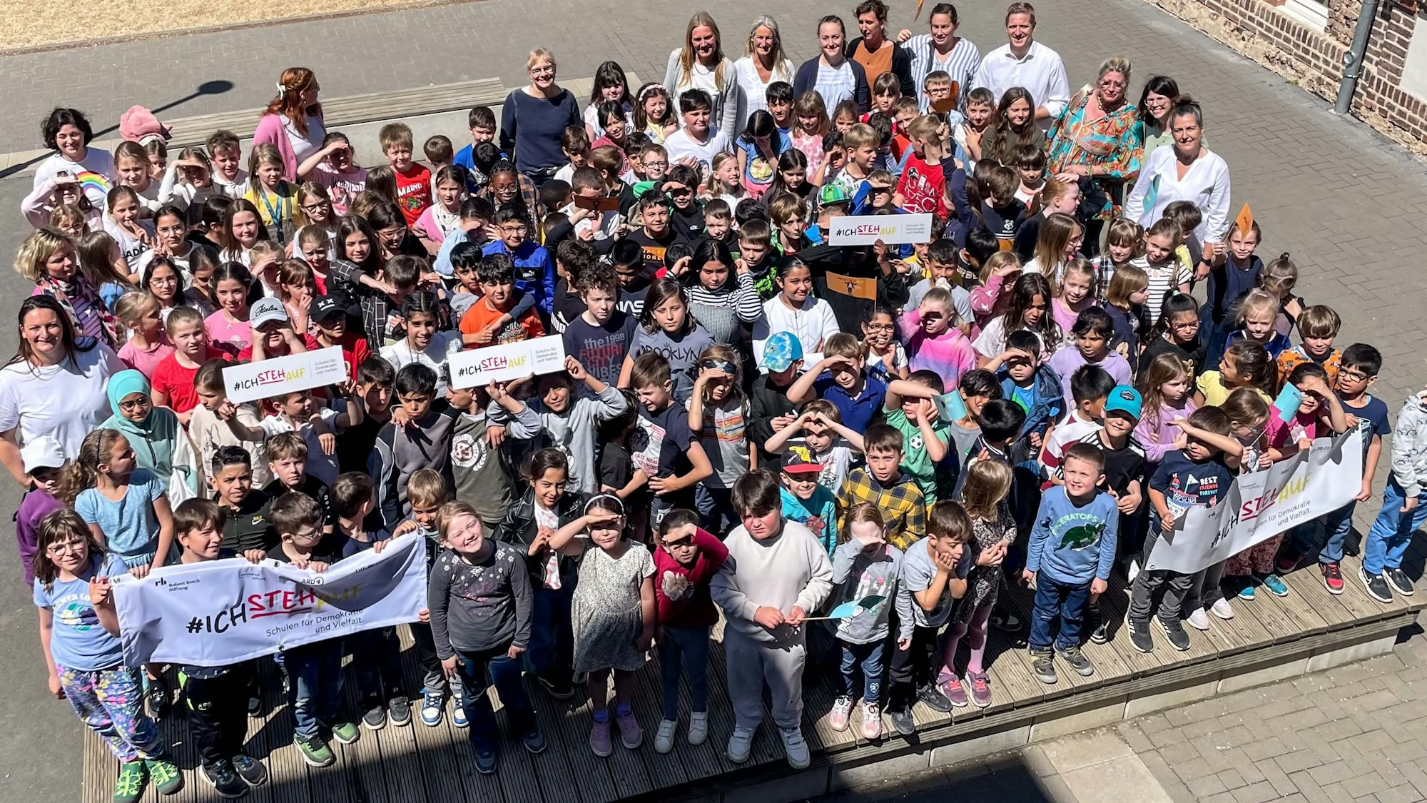 Das Bild zeigt Schüler und Lehrer der Grundschule auf dem Schulhof. Einige halten Plakate und Banner in den Händen.
