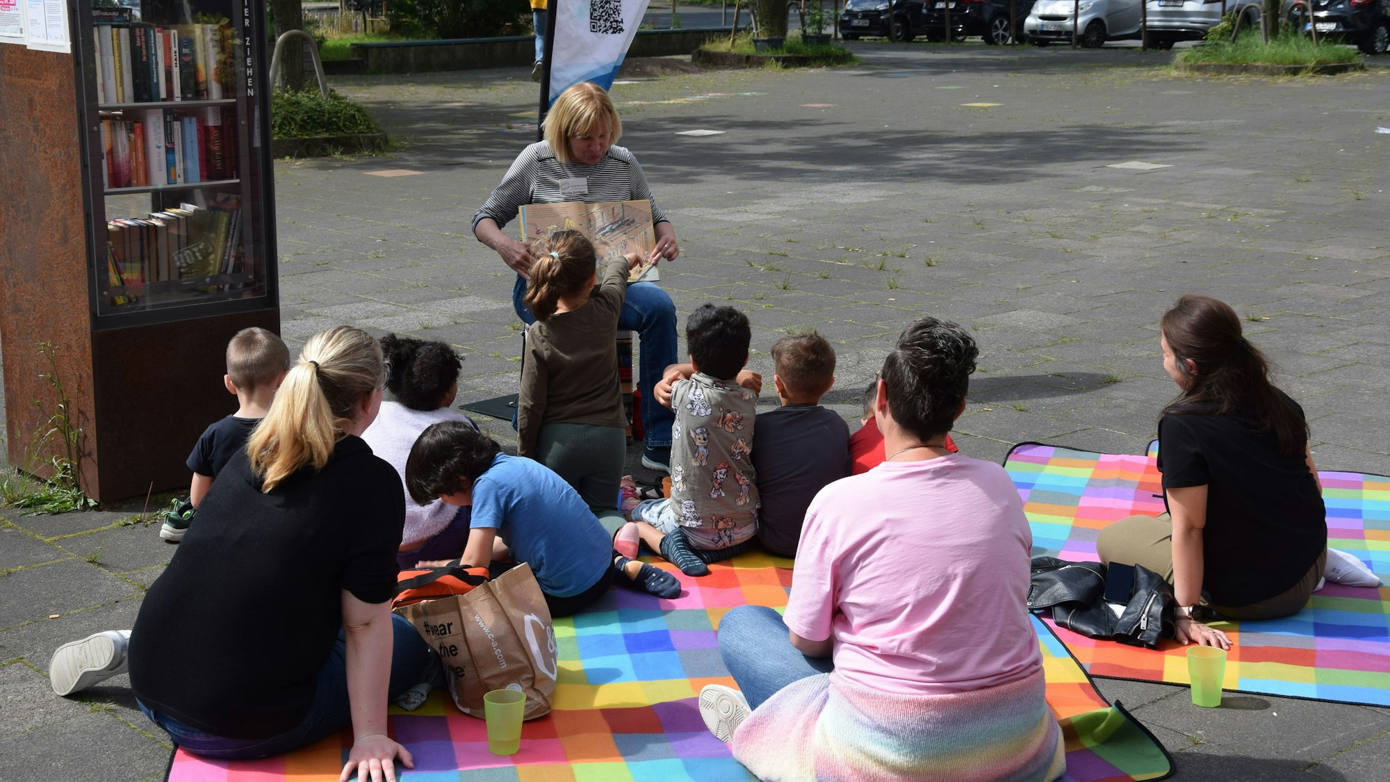 Kinder und Erwachsene sitzen auf bunten Decken auf dem Boden vor einem Bücherschrank. Eine Frau sitzt auf einem Stuhl und hält ein Buch auf ihrem Schoß hoch.